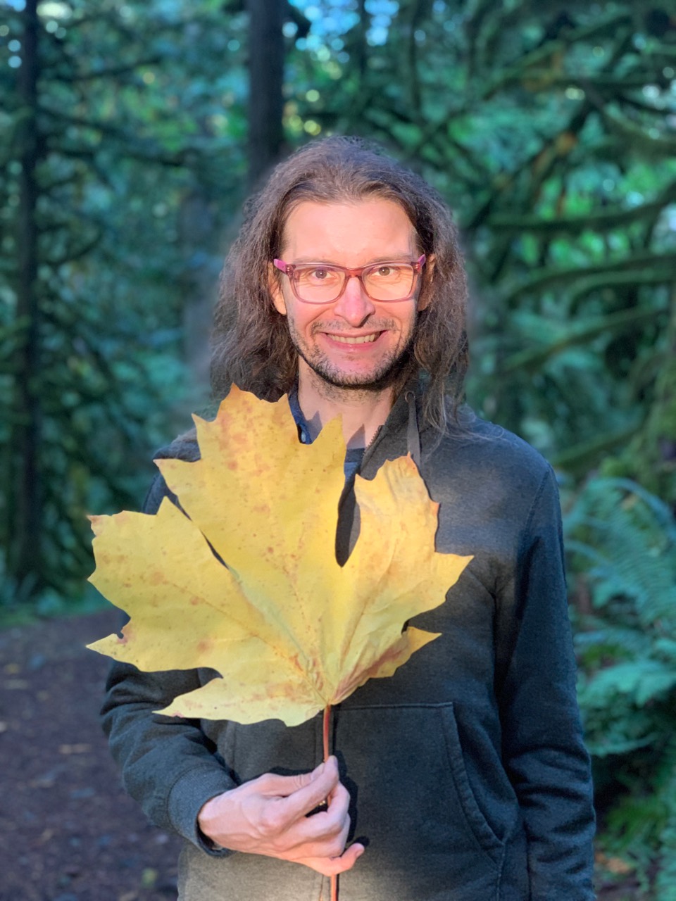 Brian Greenforest smiling in a forest while holding a large yellow maple leaf